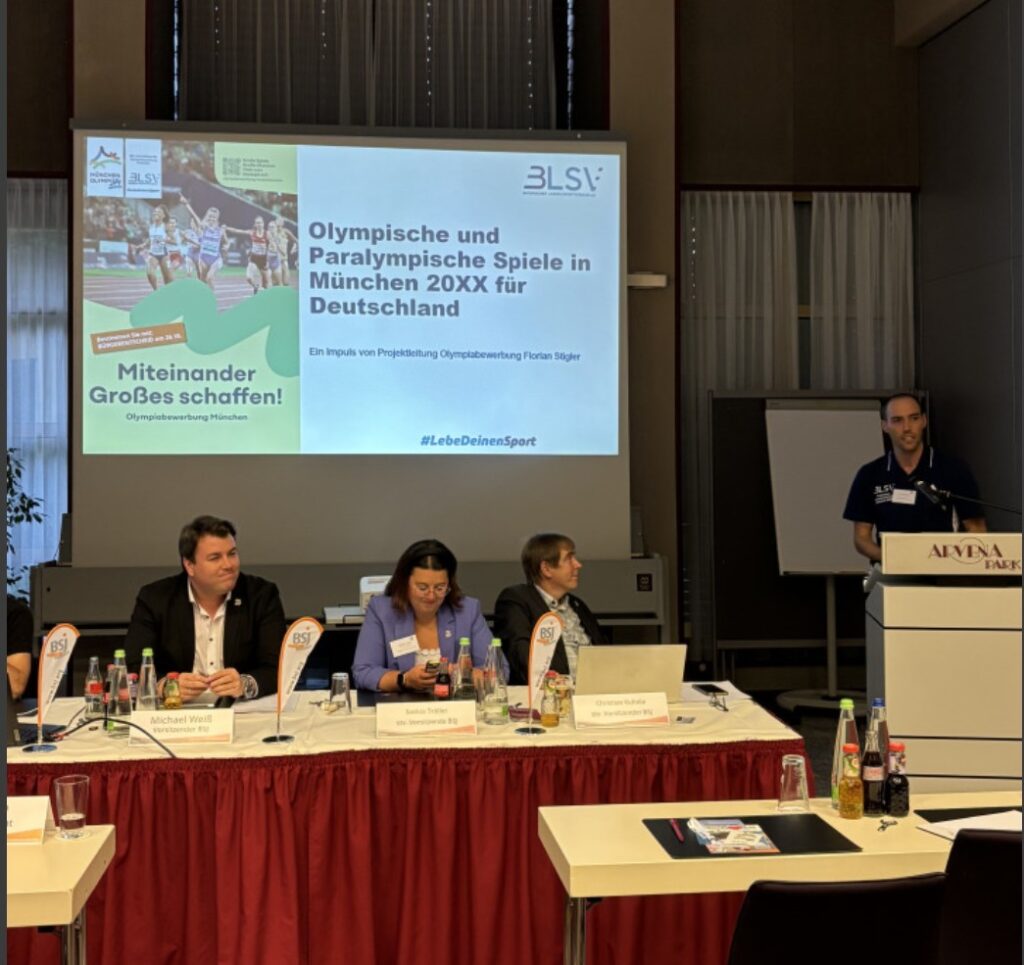 drei Personen sitzen an einem Tisch. Rechts spricht eine weitere Person am Podium. Im Hintergrund läuft eine Präsentation.