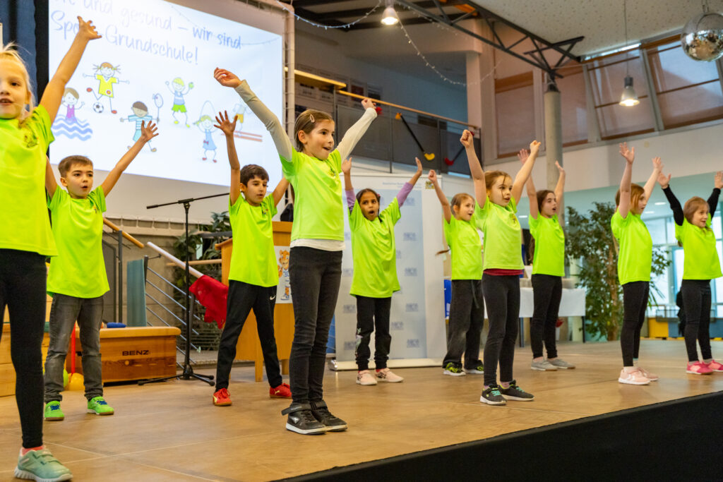 Kinder stehen auf einer Bühne und führen etwas mit hochgehobenen Händen auf.