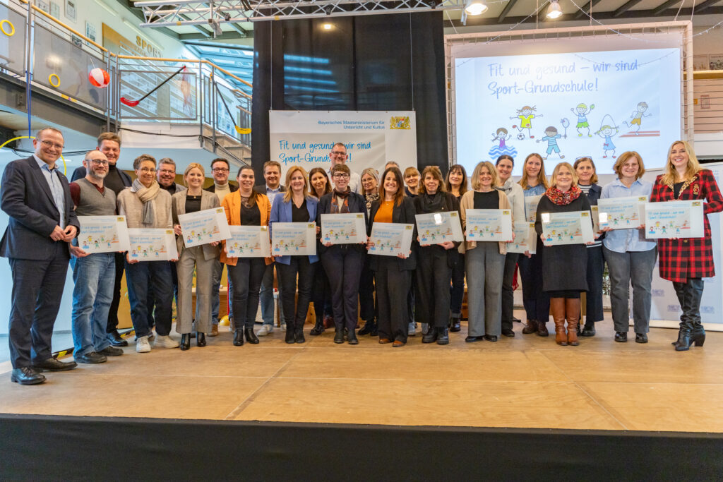 Eine Personengruppe steht zur Kamera gerichtet und viele von ihnen halten eine Urkunde zur Sportgrundschule in der Hand