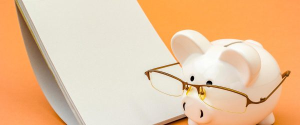 Piggy bank wearing glasses standing next to spiral notebook. Copy space.